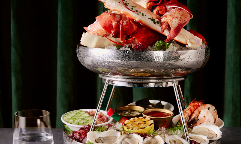 A seafood platter on a metal stand with lobster, crab legs, oysters, shrimp, sauces, and lemon, set on a gray table with a glass of water and a green curtain background.